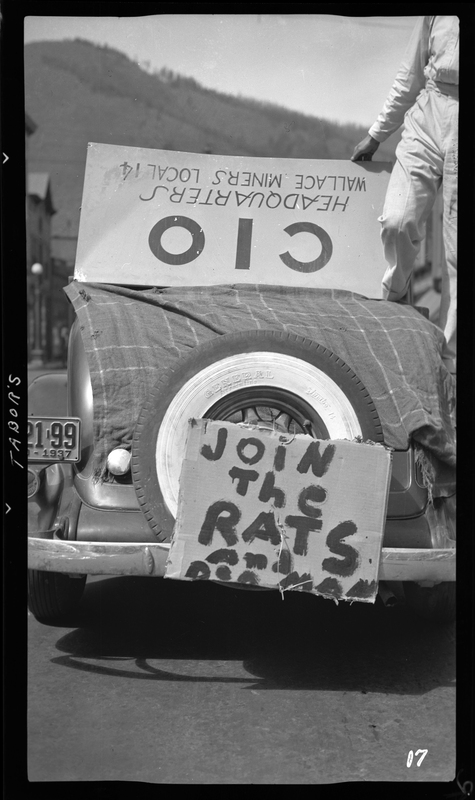 Photo of two signs on a car in Wallace, Idaho. The sign on top of the car that is being held up by a person is upside down and reads "CIO; Headquarters; Wallace Miners Local 14." The sign on the back of the car reads "Join the rats and the den man."