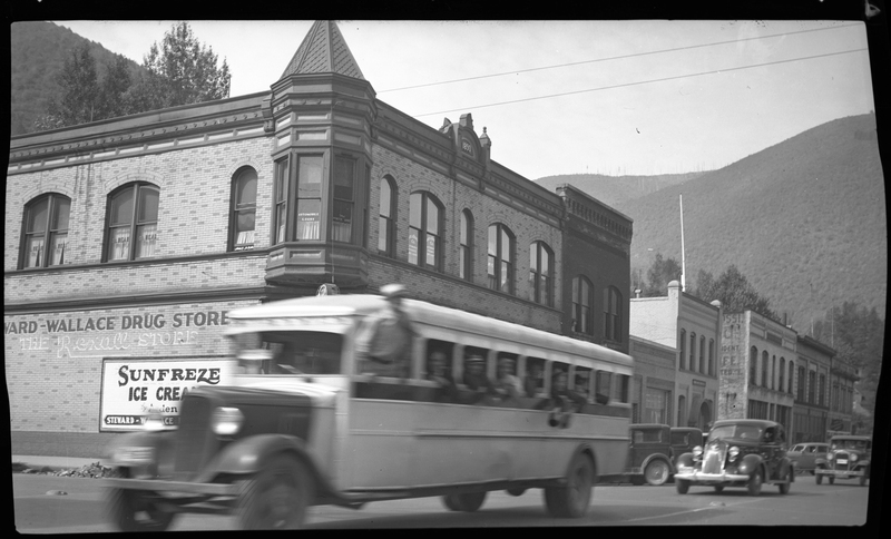 A blurry bus drives by with several people hanging out of it with cars driving behind it on the road in Wallace, Idaho. The buildings in the background are clearly visible, one of which is a Drug Store.