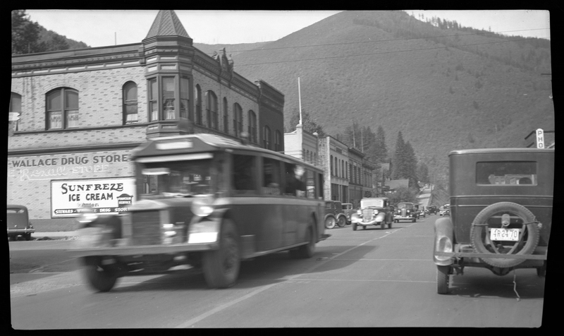 Photo of several cars driving on both sides of the road in Wallace, Idaho. The nearest vehicle driving towards the photographer is blurry from motion, but the rest of the picture is clear. The Wallace Drug Store and several other buildings are visible.