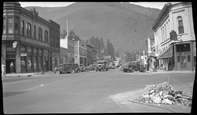 Photo of cars driving down Bank Street in Wallace, Idaho. There are piles of debris on the visible street corners, several cars parked on the sides of the road, and people walking through the shop lined street.