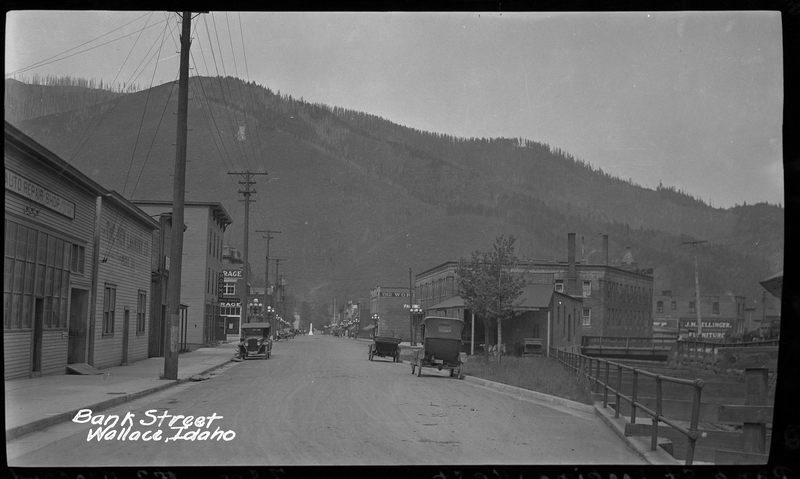 Photo of three cars parked on the side of the road on Bank Street in Wallace, Idaho. There are buildings on either side of the road, and a tree covered hill can be seen in the far background.