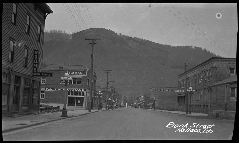 Photo of Bank Street in Wallace, Idaho taken circa 1937. There are buildings on either side of the street, and Hotel Stanley and Wallace Garage are identifiable. There are cars parked on the side of the road further down the street, and a tree covered hill in the far background.