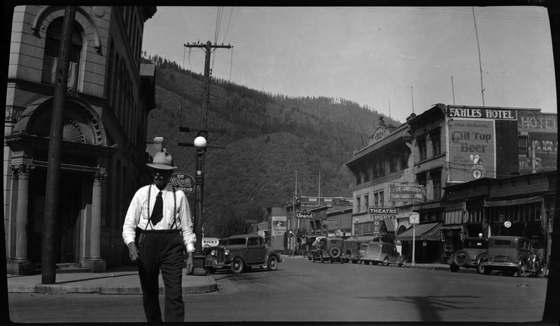Photo of an unidentified man walking across a street in Wallace, Idaho. Shops and hotels are visible along either side of the street, and several cars are parked outside.