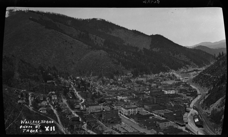 Photo taken from a high vantage point that overlooks the city of Wallace, Idaho. Several buildings and homes are visible throughout the town and the surrounding tree covered hills and mountains are easily visible around the town.