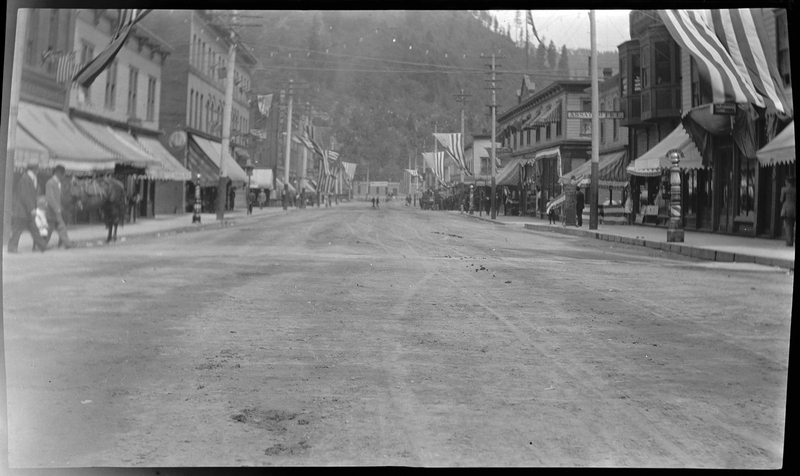 Photo of a street scene in Wallace, Idaho. American flags are hung up from telephone poles on both sides of the road, which are lined with various shops. People can be seen throughout the photo.
