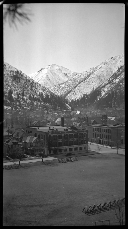 Photo taken from a high vantage point that overlooks a field and the surrounding buildings in Wallace, Idaho. The mountains that back up to the city are covered in trees and snow.