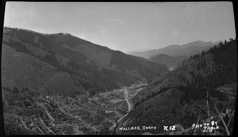 Photo taken from a high vantage point that overlooks the city of Wallace, Idaho. Several buildings and homes are visible throughout the town and the surrounding tree covered hills and mountains are easily visible around the town.