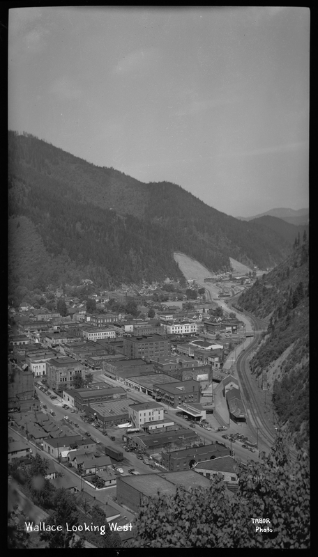 Photo taken from a high vantage point that overlooks the city of Wallace, Idaho from the east. Several buildings and homes are visible throughout the town and the surrounding tree covered hills and mountains are easily visible around the town.