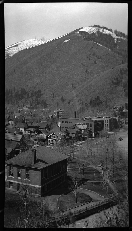 Photo taken from a high vantage point that overlooks several homes and buildings in Wallace, Idaho. The hills that back up to the city are covered in trees and snow.