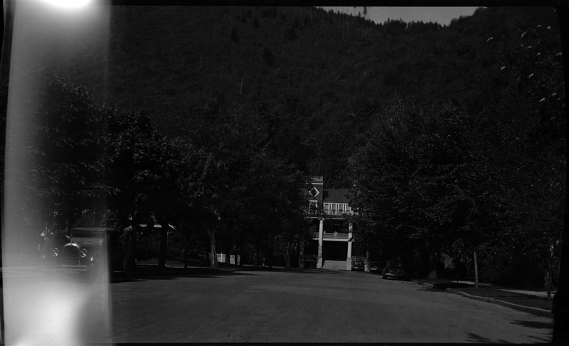 A view of Wallace Hospital in Wallace, Idaho. The left side of the negative is damaged and slightly washed out, but most of the photo is visible. Trees obscure most of the building, but the front of the building and several cars parked on the side of the road and in front of the building are visible.