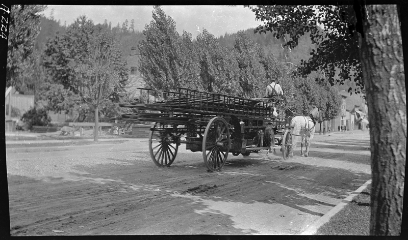 Photo of a horse drawn wagon moving down the street away from the photographer. Down the road people can be seen standing outside of buildings, but most of the sides of the road is covered in trees.