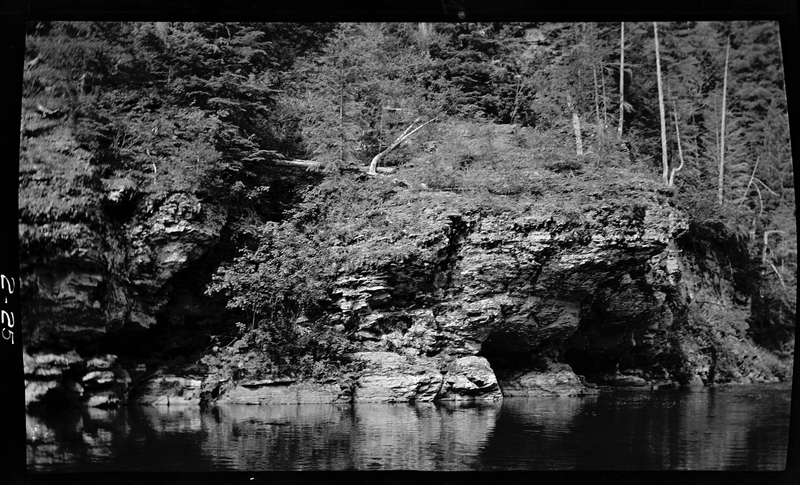 Photo of an unidentified river. Across the water is a tall rock and tree covered area and it appears there is an alcove or cave within the rock.