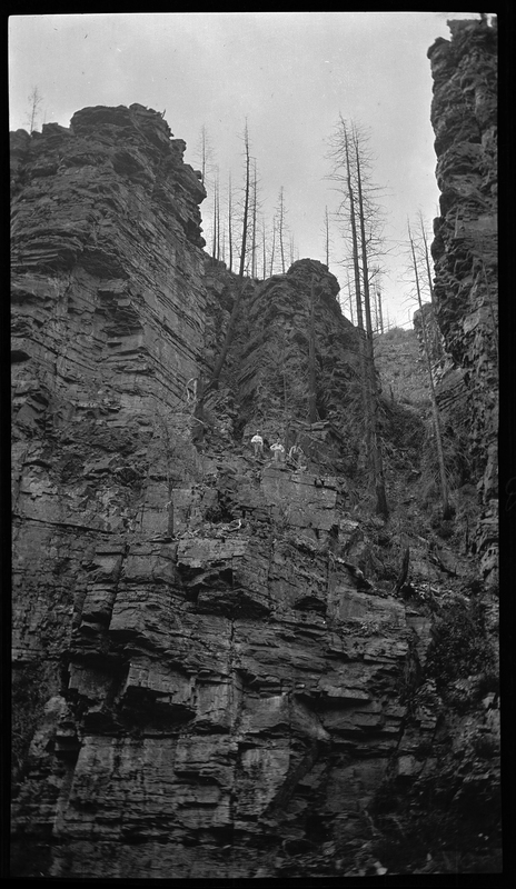 Photo of three unidentified men working on the side of a mountain. They are standing on a ledge in the middle of the photo, high above the photographer.