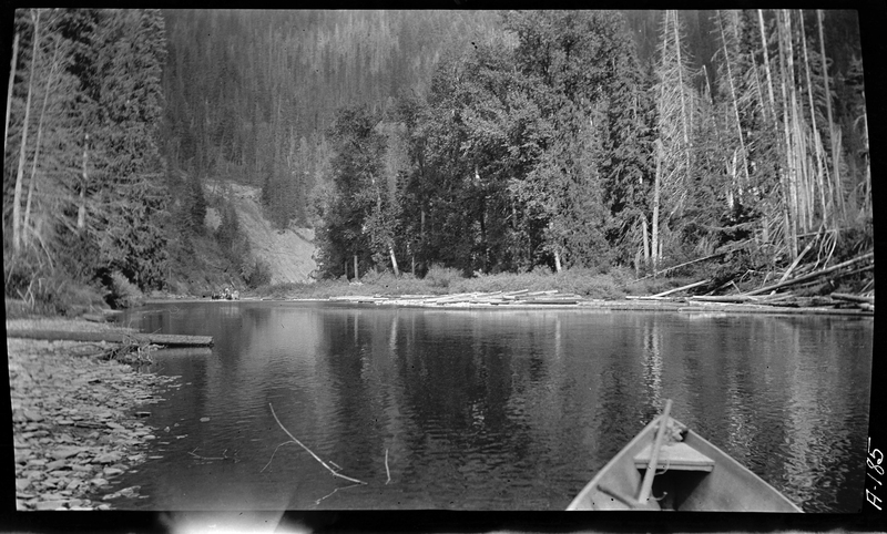 Photo of an unidentified river. The photographer is sitting in a small boat (which the front of is just visible). There is a group of people standing on the riverbank in the distance, and trees surround the river.