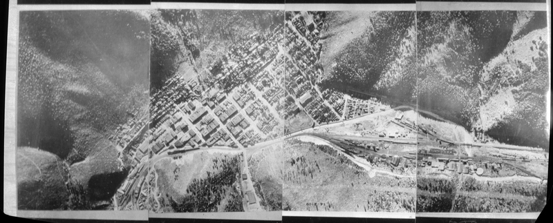 Bird eye view of a city (likely Wallace, Idaho, but unconfirmed). It appears to be four negatives taped together for the whole photo. Buildings and the surrounding landscape of mountains, hills, and trees are all visible.