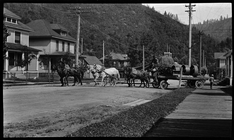 A carriage being pulled by six horses in Wallace, Idaho which was described as, "Horse carriage delivery freight east Wallace, light plant on first street." There is a man sitting on the carriage and another person walking by it. The carriage is turning onto a road lined with houses and trees.