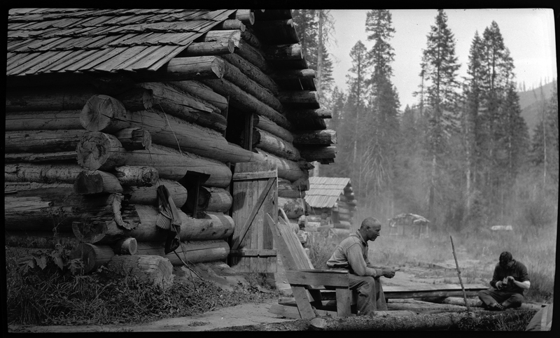 Two men sit outside of a log cabin. One man, sitting on the ground, appears to be working on something in his hands. The other man is sitting on a wooden chair. Trees can be seen in the background.