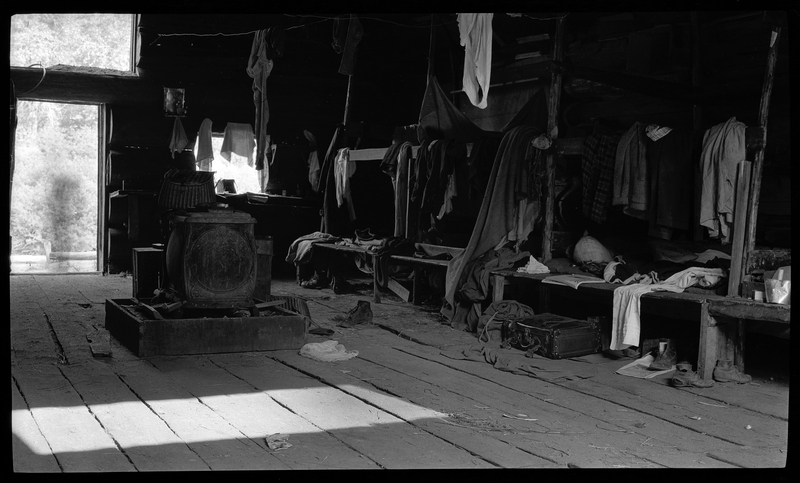 Interior photo of a log cabin. There are benches covered with clothes and other personal items along the wall, and a piece of machinery in the middle of the room.