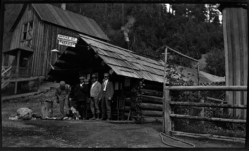 Photo of four unidentified men and a dog standing just outside of a log built building with signs labelling it as "Office of Paragon Consolidated Mining Co." and "Paragon Post Office." There is another building behind the Post Office, and trees directly behind both buildings. 