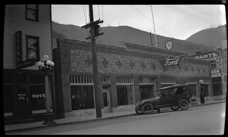 Photo of a newly built Ford Garage dealership in what appears to be Wallace, Idaho. There is a sign on the building that reads "Ford; Authorized Dealers; C. E. Haines Co., Inc." Outside of the building there is a car parked on the side of the street, and what is likely a gas pump.