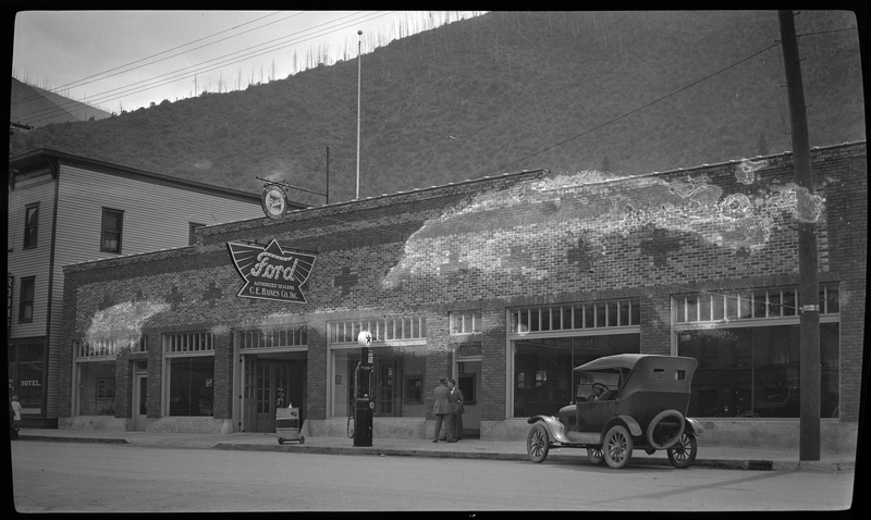 Photo of a newly built Ford Garage dealership in what appears to be Wallace, Idaho. There is a sign on the building that reads "Ford; Authorized Dealers; C. E. Haines Co., Inc." Outside of the building there is a car parked on the side of the street, and what is likely a gas pump, as well as two men standing outside of the door.