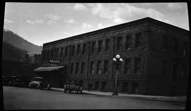 Photo of the Pacific Hotel in Wallace, Idaho. The photo is grainy and dark, but some detail can be made out. There are cars parked on the road in front of the building, as well as a man walking down the sidewalk.