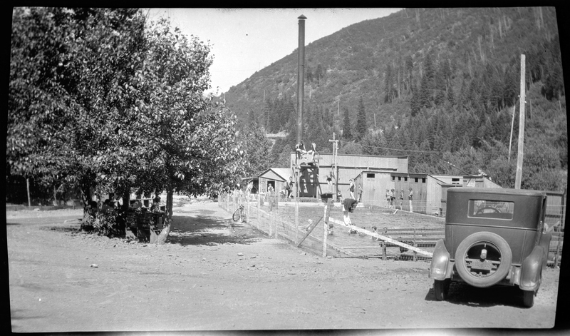 Photo of an in ground swimming pool in Wallace, Idaho. Labelled at the first swimming pool, several people can be seen swimming or standing around the pool, which is fenced in. There is a car parked outside of the area, which stands between the photographer and the pool itself. There are trees seen next to the pool and on the hills behind it.