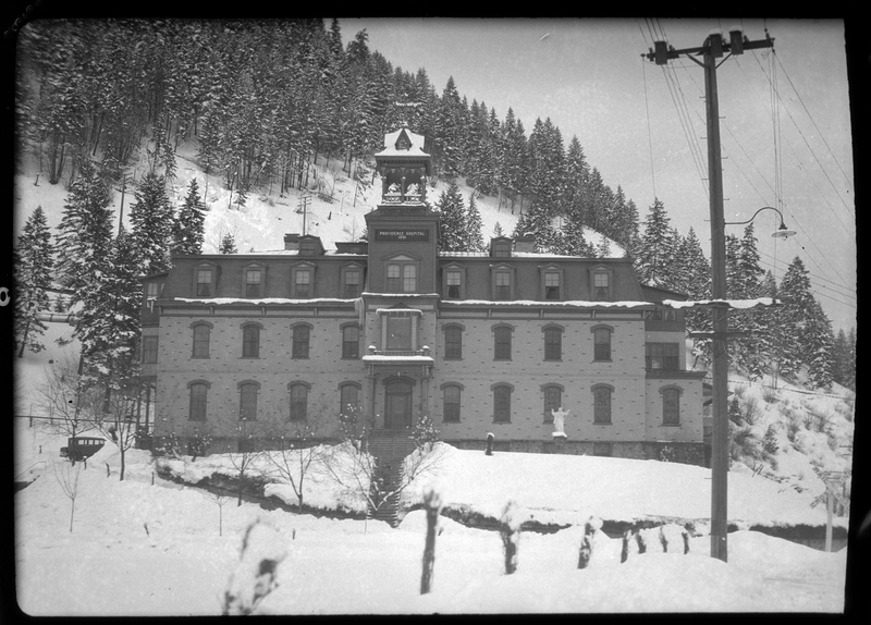 Photo of the Providence Hospital in Wallace, Idaho in the snow. The sign near the top of the three story building reads, "Providence Hospital 1891." The ground surrounding the building, the roof of the building, and the tree covered hill behind the hospital are all covered in a heavy layer of snow. 