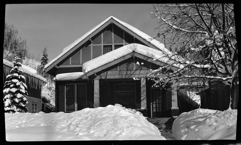 Photo of George Edminston's House, 100 block on Cedar Street, in Wallace, Idaho. The roof of the house, surrounding ground, and trees are all covered in a heavy layer of snow.