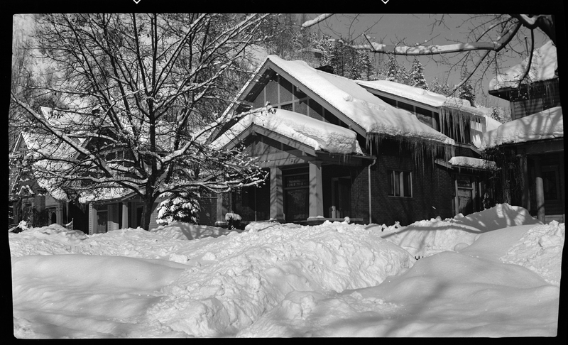 Photo of George Edminston's House, 100 block on Cedar Street, in Wallace, Idaho. The roof of the house, surrounding ground, and trees are all covered in a heavy layer of snow. Large icicles can be seen hanging off the roof on the side of the house.