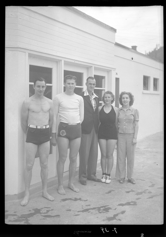 Photo of three men and two women at the Wallace, Idaho swimming pool. Two of the men and one of the women are dressed to swim, and it appears that at least one of the men is a lifeguard. The other man and woman are dressed nicely. They are standing outside of a building and are posing for the photo.
