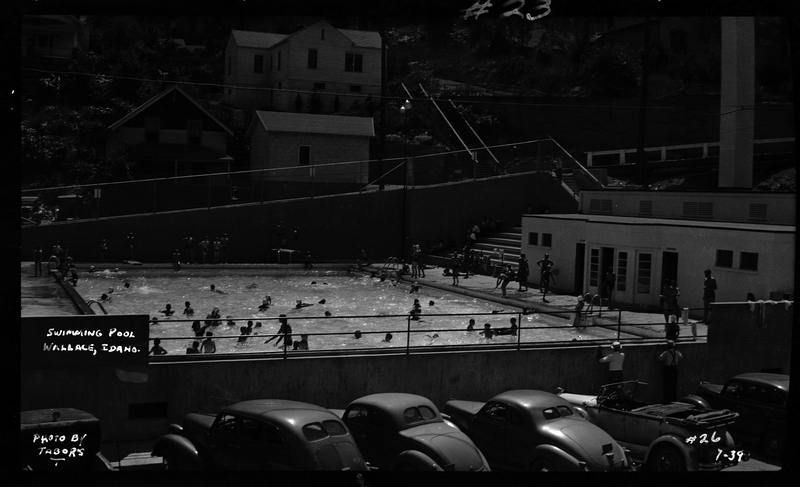 Photo of a large number of people swimming in the Wallace, Idaho Swimming Pool. There are people on the concrete outside of the pool, but a majority of people are actively swimming. There are several cars parked outside of the blocked off pool.