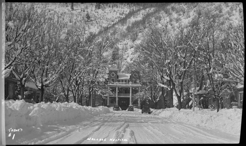 Photo of the Wallace, Idaho hospital in the snow. The building appears to be at least three stories tall. The photographer is standing down the road, facing the hospital, which is partially obscured by trees on both sides of the road. There are also three cars, one of which is parked on the side of the road and the other two parked in front of the hospital. The ground, trees, and roof of the hospital are all covered in a heavy layer of snow.