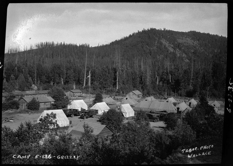 Photo described as "Civilian conservation corps camps during the 1930s, Shoshone County," with Camp-F-136-Grizzly written on the negative. Several buildings and tents are set up in the clearing of a forest, and several trucks are visible.