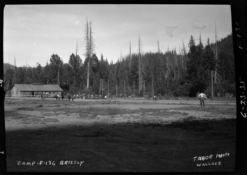 Photo described as "Civilian conservation corps camps during the 1930s, Shoshone County," with Camp-F-136-Grizzly written on the negative. Several people can be seen standing in the distance in a cleared out area of a forest. There is a building off to one side of the clearing.