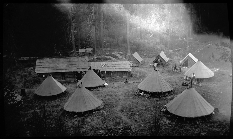 Photo described as "Civilian conservation corps camps during the 1930s, Shoshone County." There are several tents surrounding a single building in a clearing in a forest. Several people sit and stand among the tents.