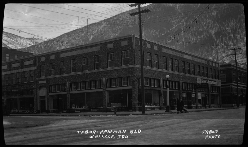 Photo of the Tabor-Pfirman building in Wallace, Idaho. The photographer stands opposite of the corner of the building, and two people can be seen crossing the street. There is a sign on one side of the building that reads "Stone's Food Stores."