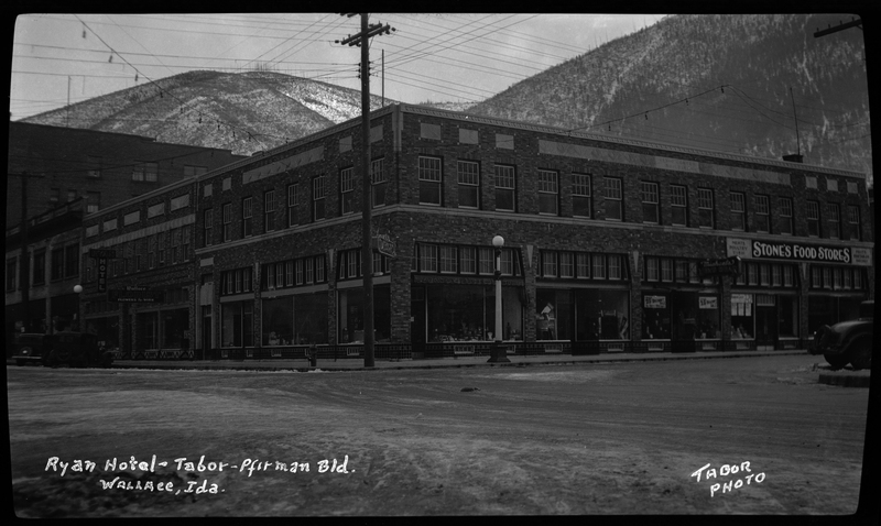 Photo of the Tabor-Pfirman building and the Ryan Hotel in Wallace, Idaho. The photographer stands opposite of the corner of the building and snow is visible on the road between them. There is a sign on one side of the building that reads "Stone's Food Stores," and the sign for the Ryan Hotel can be seen on the other side of the building.