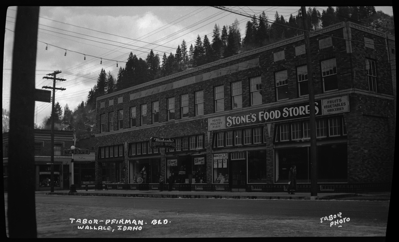 Photo of the Tabor-Pfirman building in Wallace, Idaho. The photographer stands across the street from the building. There is a sign on one side of the building that reads "Stone's Food Stores," as well as other signs advertising food, one that reads "Michaels Men's Wear," and one on the corner of the building that reads "Tabor's."