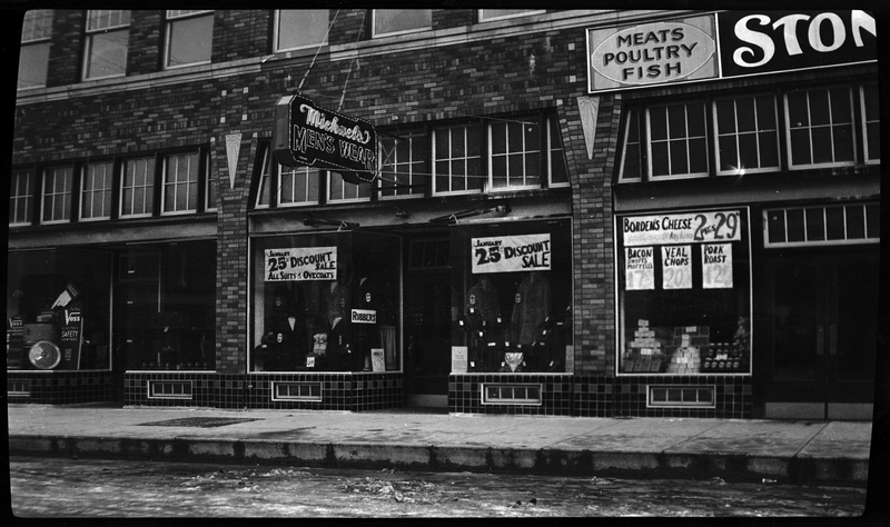 Photo of Michael's Men's Wear store in Wallace, Idaho. There is another store next to it, but the sign is cut off in the picture. There are advertising signs in the windows, and what appears to be water in the street.