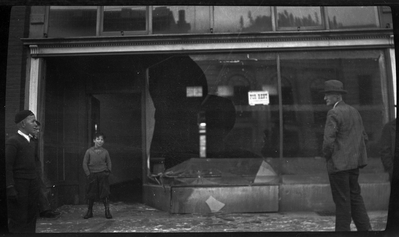 Photo of several children and a man who are standing outside of a building. The building as two large windows, one of which had been broken. There is glass covering the ground in front of the building.