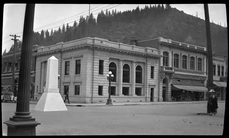 Photo of the First National Bank and the Mullan Monument, the latter of which stands in the middle of an intersection. There are a few people walking on the sidewalks, and a car parked on the side of the road next to the bank.
