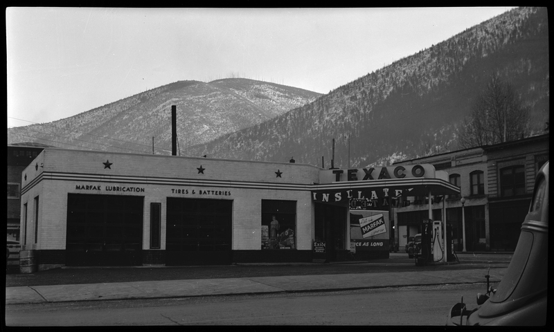 Photo of a Texaco gas station in Wallace, Idaho. The gas station has two garages, one with "Marfak Lubrication" above it and the other with "Tires & Batteries" above it. There are signs advertising for Marfak, insulation, and batteries around the visible parts of the building.