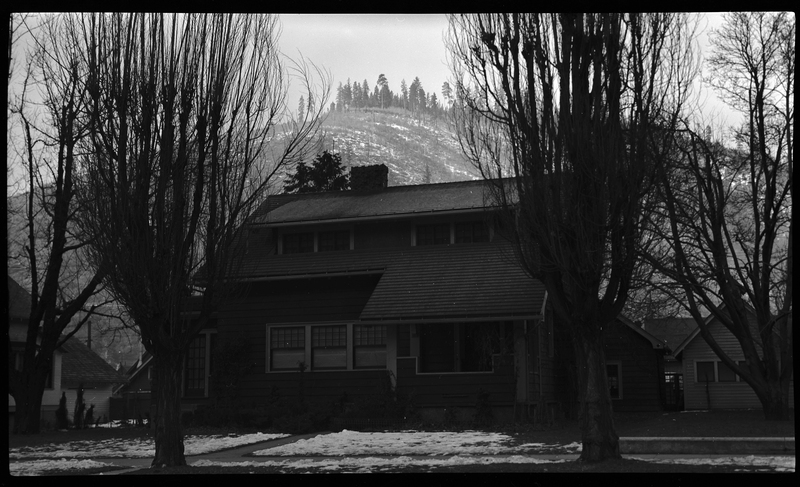 Photo of a home in Wallace, Idaho. The photo was taken from across the street, showing the front of the house. There are some partially melted piles of snow on the ground, and two large trees in the front yard.