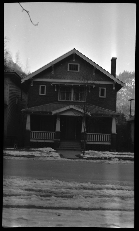 Photo of a home in Wallace, Idaho. The photo was taken from across the street, showing the front of the house. There are some partially melted piles of snow on the ground, and a tree in the front yard.