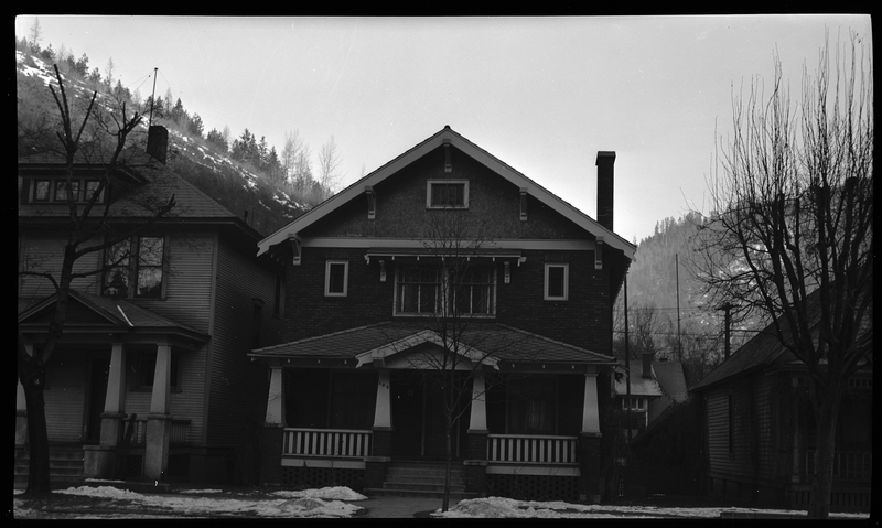 Photo of a home in Wallace, Idaho. The photo was taken from across the street, showing the front of the house. There are some partially melted piles of snow on the ground, and a tree in the front yard.