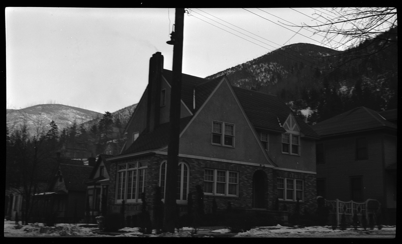 Photo of a home in Wallace, Idaho. The house sits on a corner and the photo was taken across the street from that corner, showing the front and side of the house. There are some partially melted piles of snow on the ground.