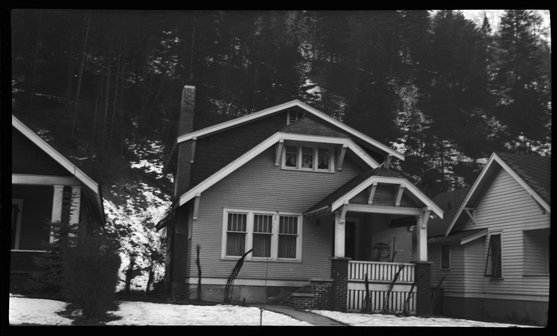 Photo of a home in Wallace, Idaho. The photo was taken from across the street, showing the front of the house. There is a small layer of snow on the ground surrounding the house, but the sidewalk is clear.