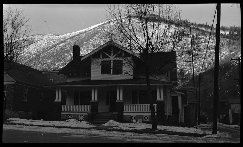 Photo of a home in Wallace, Idaho. The house sits on the corner of a street and photo was taken from across the street, showing the front of the house. There is a tree in the front yard and a small layer of snow on the ground surrounding the house, but the sidewalk is clear.
