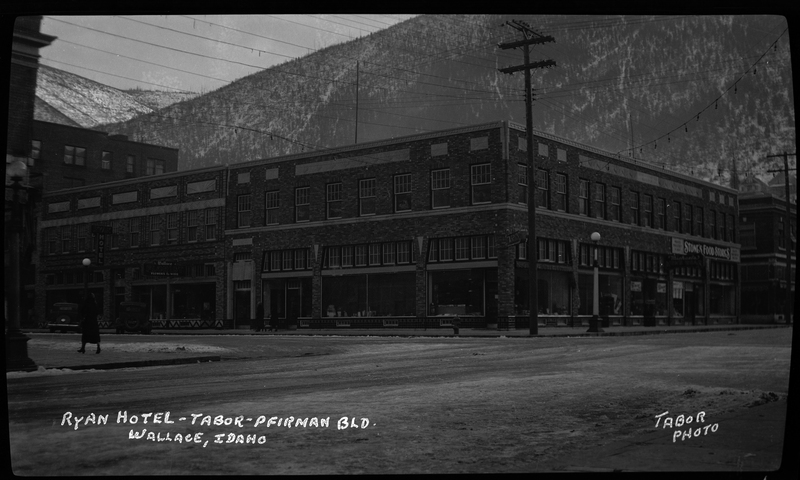 Photo of the Tabor-Pfirman building and the Ryan Hotel in Wallace, Idaho. The photographer stands opposite of the corner of the building and snow is visible on the road between them. There is a sign on one side of the building that reads "Stone's Food Stores," and the sign for the Ryan Hotel can be seen on the other side of the building.